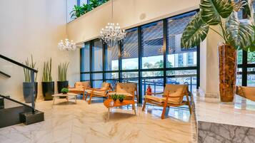 A lobby with a marble floor, wooden chairs, a chandelier, large windows, and potted plants.