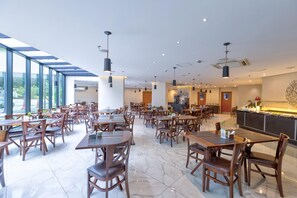Un restaurante espacioso con techos altos, lámparas colgantes modernas y un amplio muro de ventanas que proporciona luz natural.
