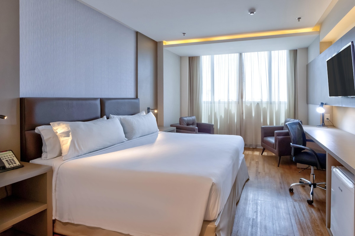 Habitación de hotel con una cama grande, un escritorio con silla, televisor y ventana con cortinas.