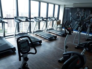 Un gimnasio con cintas de correr, elíptica y bicicleta estática, ventanas amplias con vista a la ciudad y un cartel en la pared.