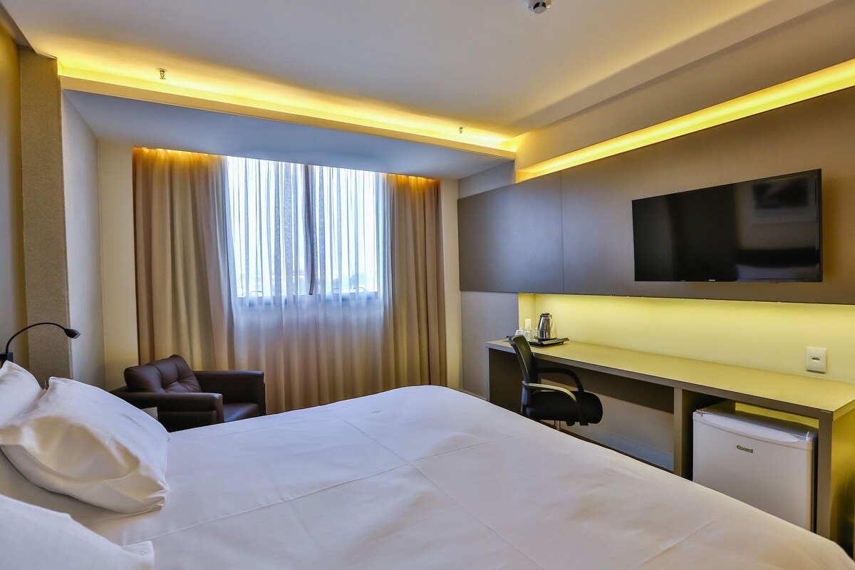 Habitación de hotel con cama, escritorio, silla, televisor y refrigerador pequeño.