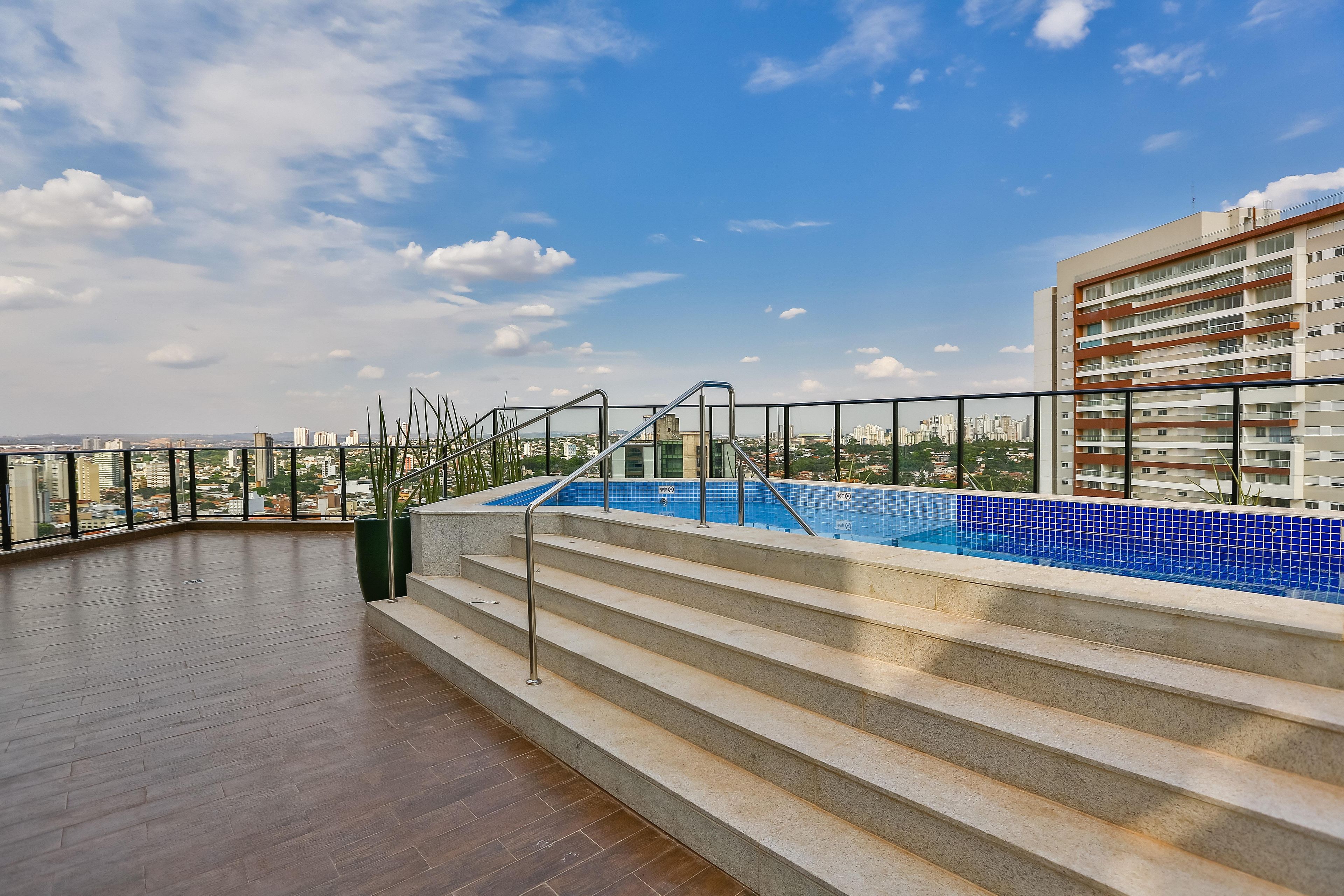 A rooftop pool area with steps leading to a blue-tiled pool, surrounded by a railing and a view of a cityscape.