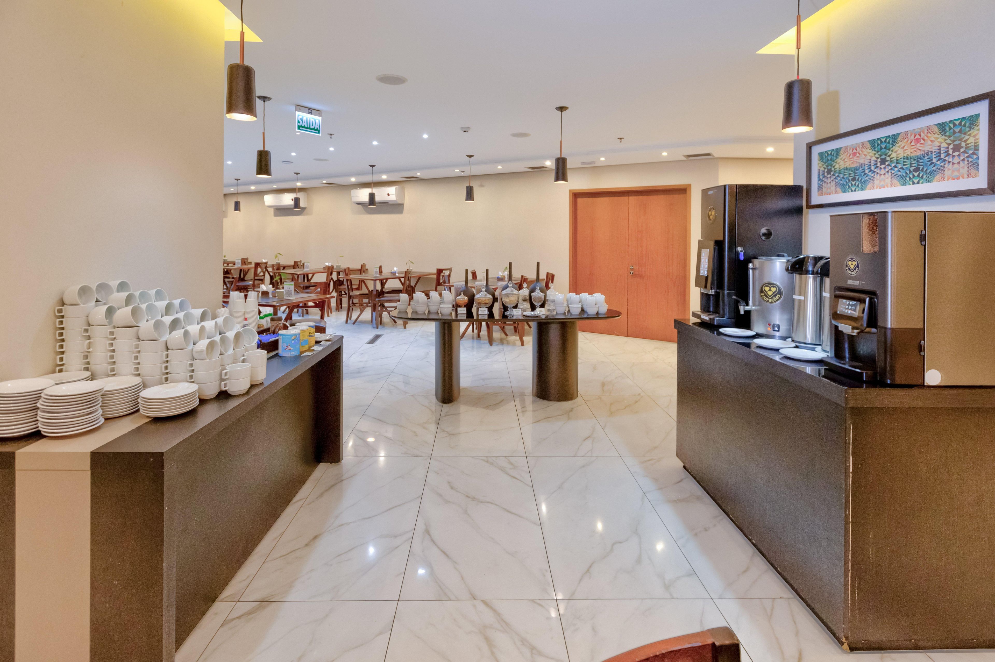 A hotel lobby with a marble floor, a central dining table surrounded by chairs, a coffee station, and a framed artwork on the wall.