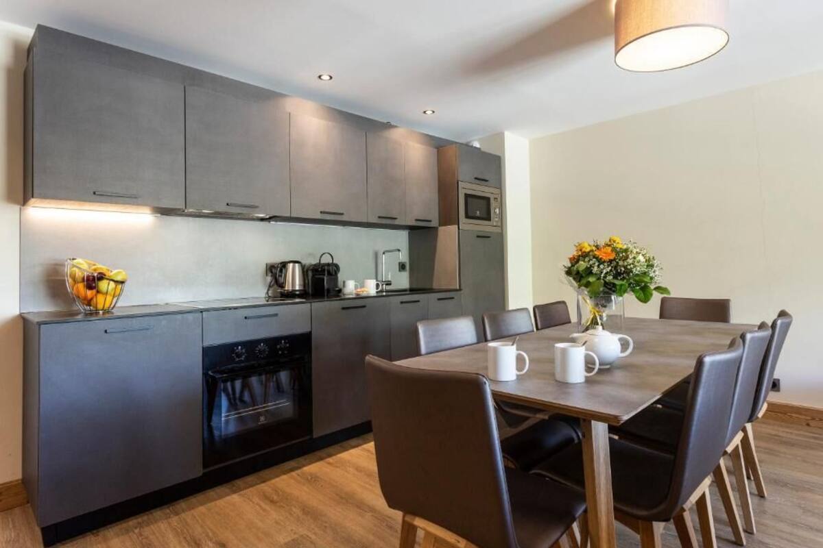 A modern kitchen with dark cabinets, a built-in oven, and a dining area with a table and chairs.