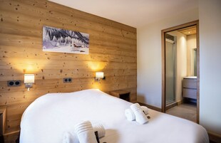 Camera d'albergo con parete in legno, un letto con lenzuola bianche, due luci a parete, un quadro che raffigura un paesaggio innevato e un bagno visibile attraverso una porta in vetro.