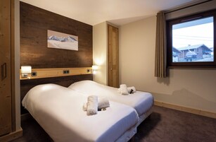 A hotel room with two beds, wooden paneling, a mountain painting, a window with a view, and a lamp on the nightstand.