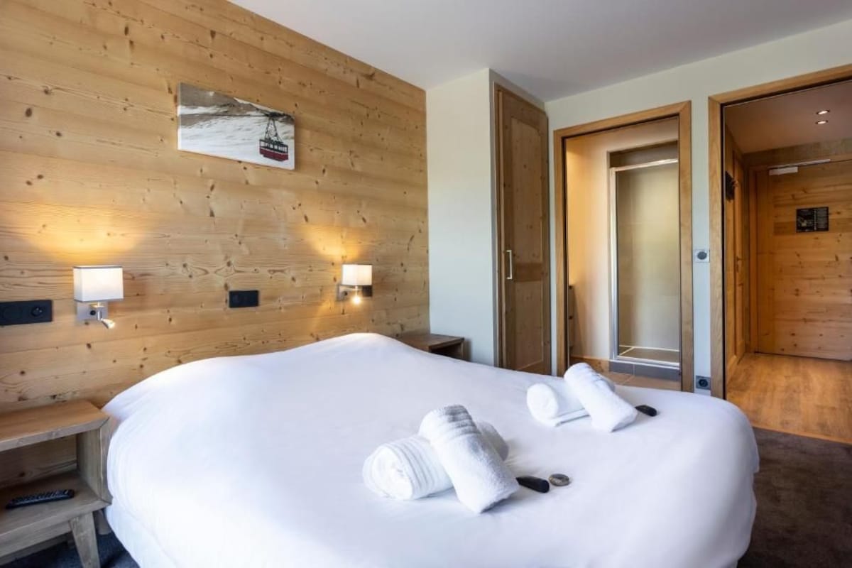 A hotel room with a wooden wall, a bed with white linens, two wall-mounted lamps, and a door leading to another room.