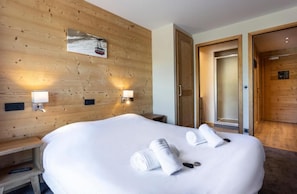 A hotel room with a wooden wall, a bed with white linens, two wall-mounted lamps, and a door leading to another room. - Residence Daria-i Nor (Huez)