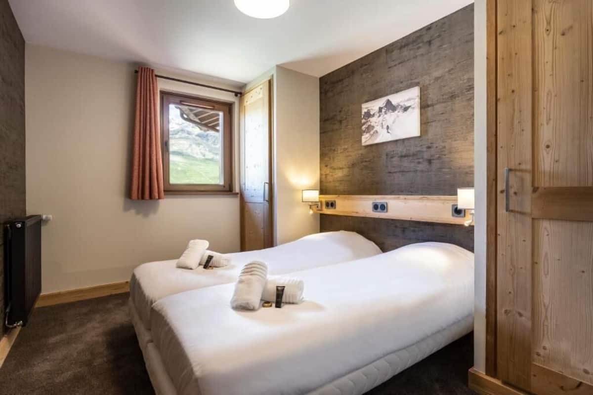 A hotel room with two beds, a wooden wall, a window with curtains, a radiator, and a picture on the wall.