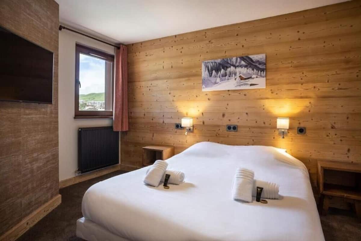 A hotel room with a wooden wall, a bed with white linens, two wall-mounted lamps, a window with a view, and a painting above the bed.