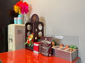 Uma mesa com dispensador de água, um vaso de flores, uma máquina de café, um dispensador de lanches e uma placa indicando alimentos e bebidas para presente.