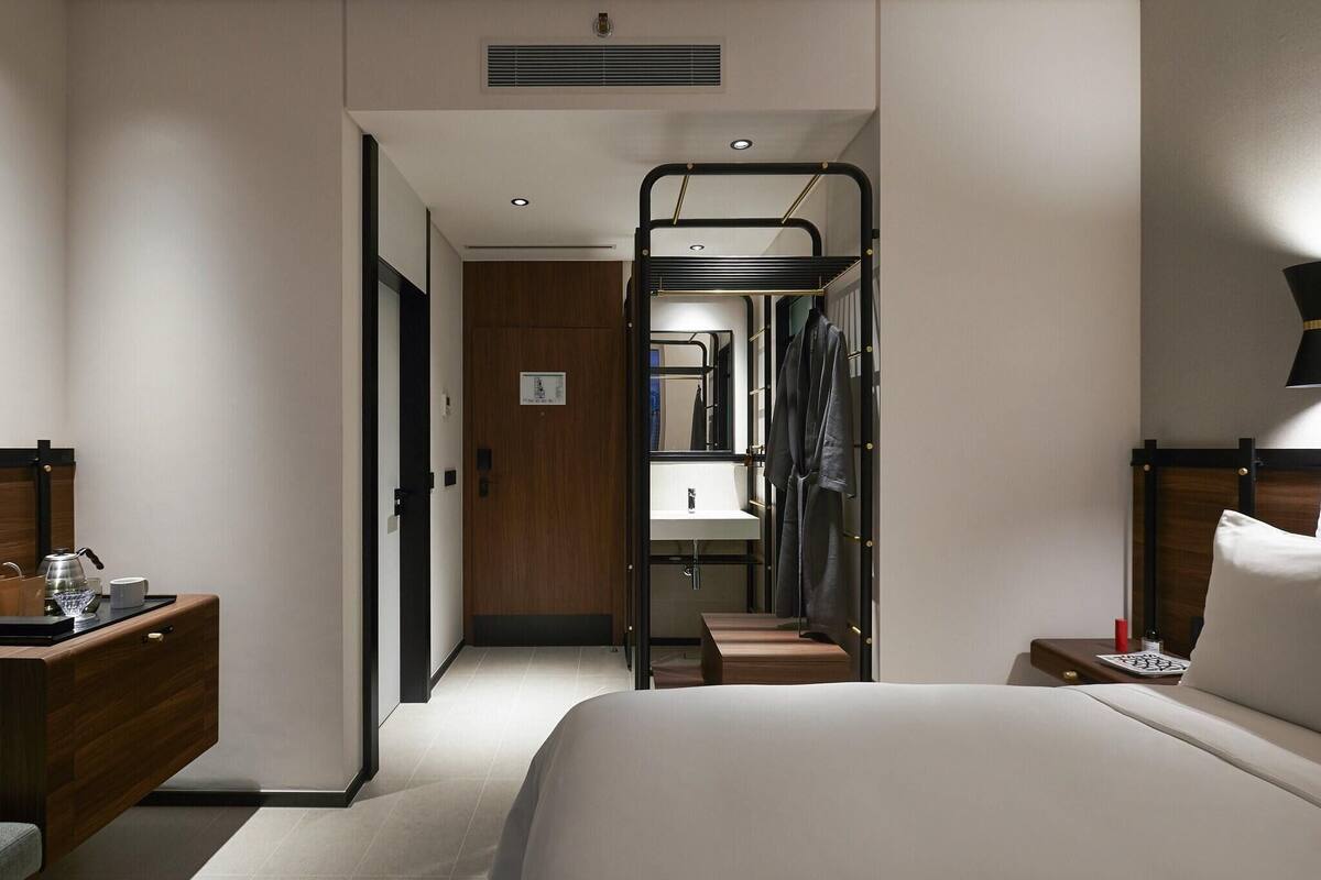 Ett hotellrum med en våningssäng, ett sängbord med en lampa, ett litet bord, en byrå i trä och ett badrum med handfat och spegel.