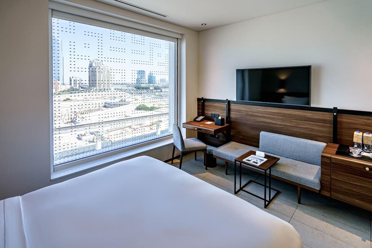 Ett hotellrum med en säng, ett skrivbord, en stol, en TV och ett stort fönster med utsikt över staden.
