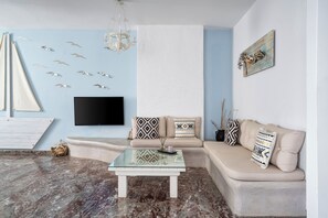 A living room with a beige sofa, a glass coffee table, and a wall-mounted TV. - Iris Boutique Hotel (Paros)