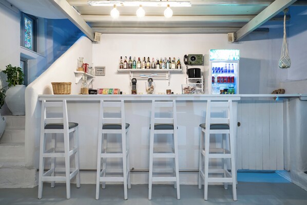 A bar area with white bar stools, a shelf with bottles, a coffee machine, and a refrigerator. - Iris Boutique Hotel (Paros)