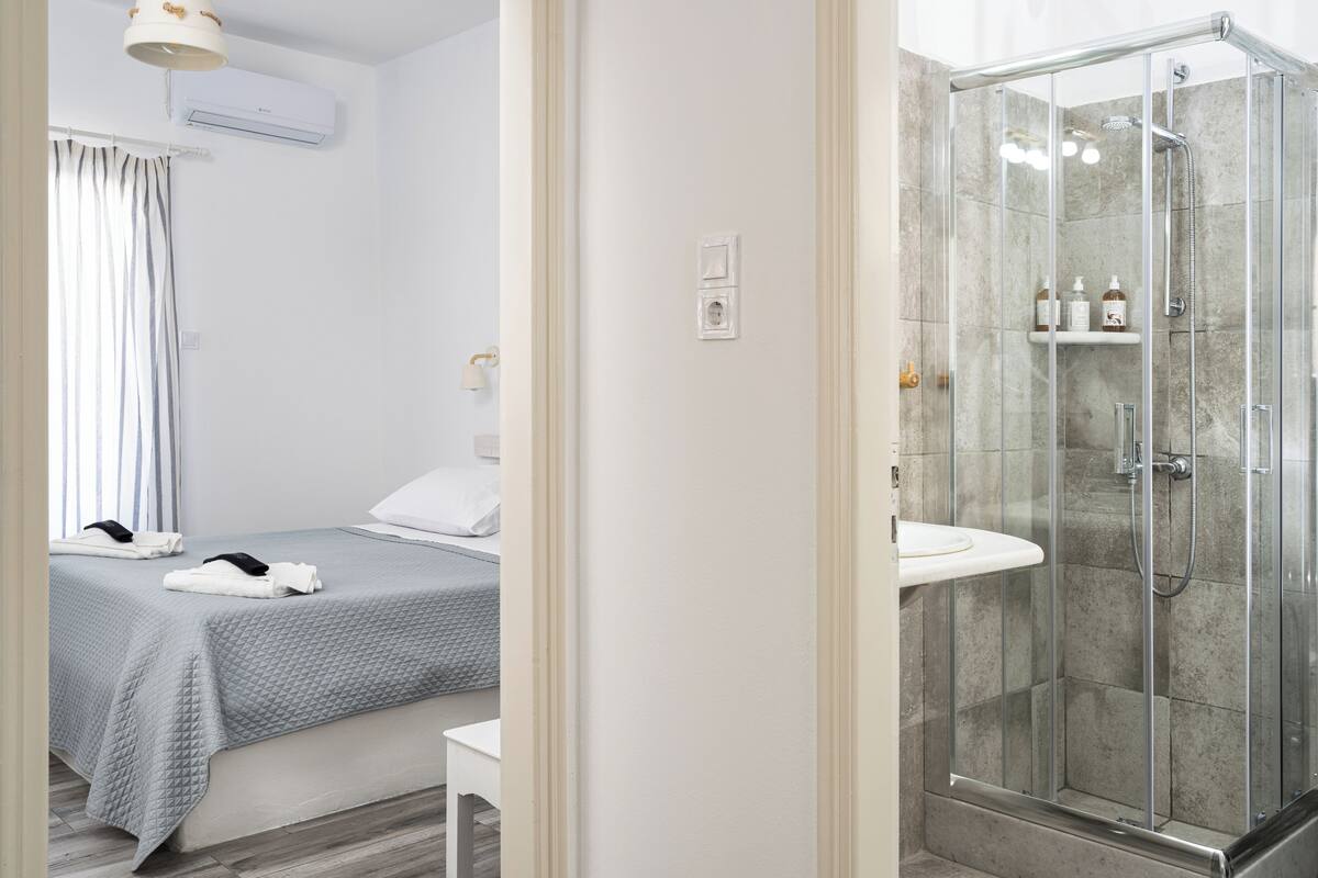 A hotel room with a bed, a bedside table, a shower with a glass enclosure, and a bathroom sink.