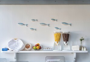 A breakfast bar with a white cloth, a basket of eggs, a bowl of fruit, two coffee dispensers, and a vase with flowers. - Iris Boutique Hotel (Paros)