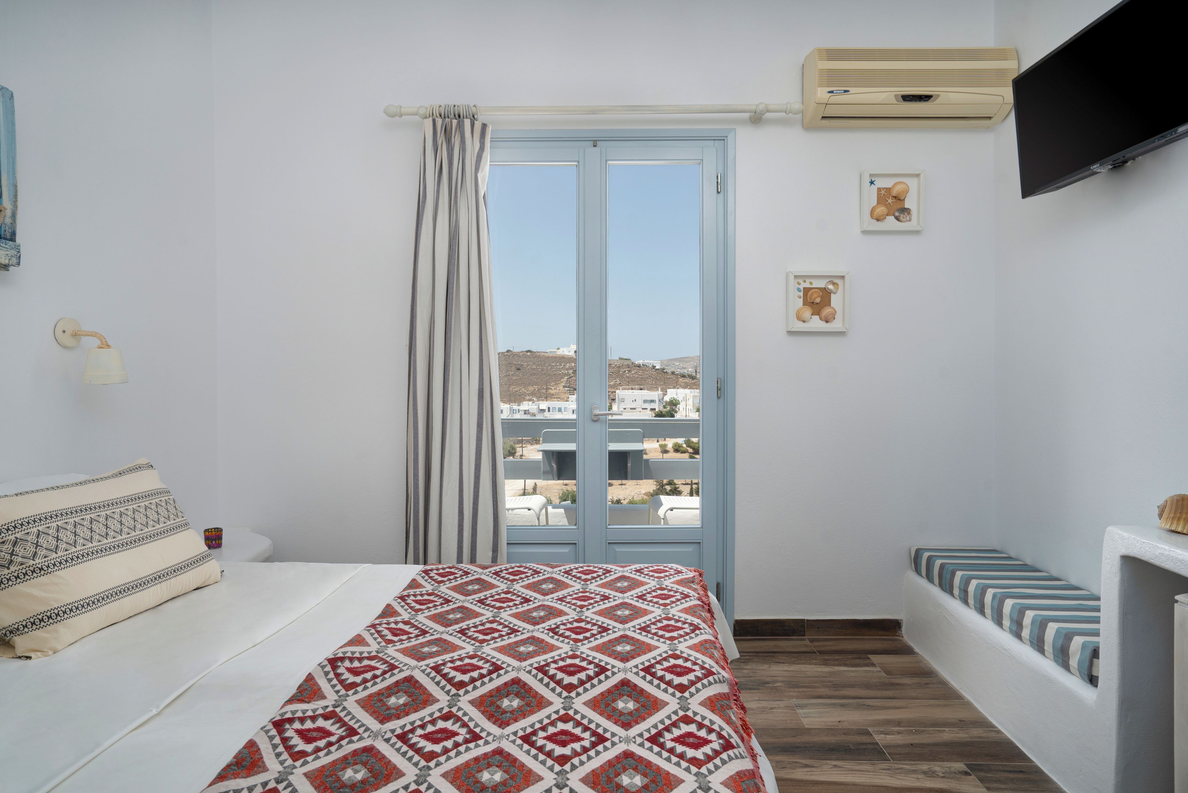 A bedroom with a bed, a window with curtains, a mounted TV, and a small table with a lamp.