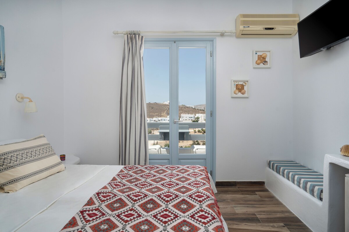 A bedroom with a bed, a window with curtains, a mounted TV, and a small table with a lamp.