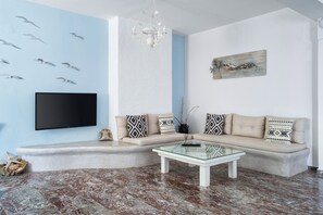 A living room with a beige sofa, patterned cushions, a glass coffee table, a flat-screen TV, and a wall-mounted artwork. - Iris Boutique Hotel (Paros)