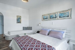 A bedroom with a large bed, two bedside tables, a nightstand, a white sofa, and a wall-mounted telephone. - Iris Boutique Hotel (Paros)
