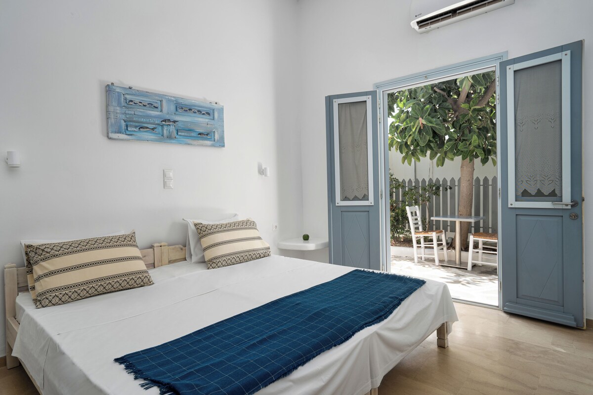 A bedroom with a bed, two pillows, a blue blanket, a wall-mounted artwork, and a door leading to an outdoor area with a tree and a white fence.