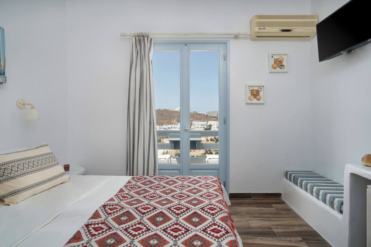 A bedroom with a bed, a window with curtains, a mounted TV, and a small table with a lamp.