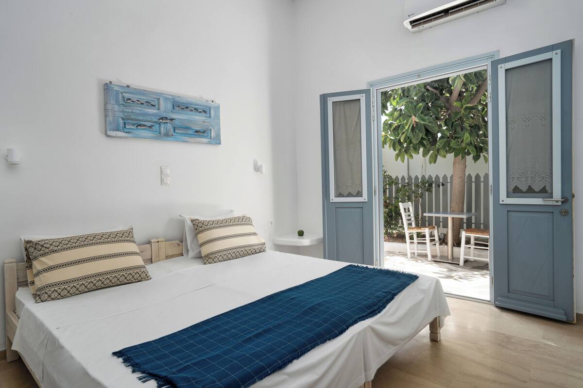 A bedroom with a bed, two pillows, a blue blanket, a wall-mounted artwork, and a door leading to an outdoor area with a tree and a white fence.