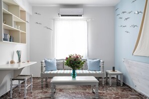A room with a white sofa, a small table, a bookshelf, and a window with curtains. - Iris Boutique Hotel (Paros)