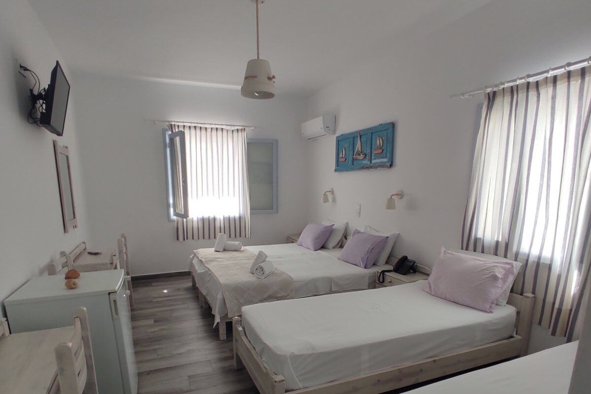 A room with two beds, a desk, a chair, a window with curtains, a wall-mounted TV, and a ceiling fan.