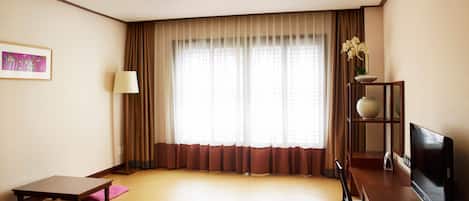 A hotel room with a large window, a flat-screen TV, a wooden desk, and a small shelf with a vase.