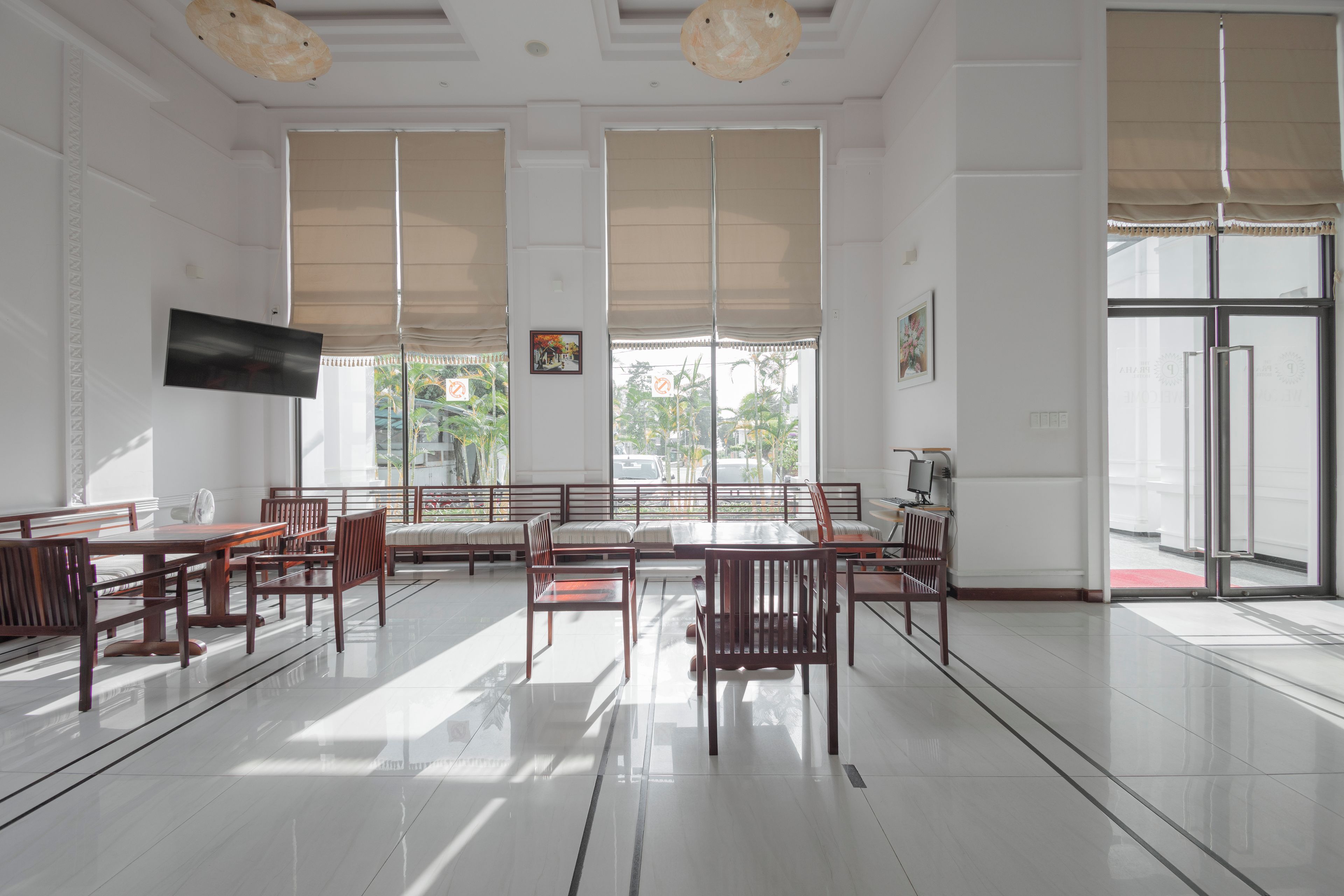 A spacious room with a large dining table, multiple chairs, a flat-screen TV mounted on the wall, and large windows with blinds.