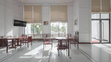 A spacious room with a large dining table, multiple chairs, a flat-screen TV mounted on the wall, and large windows with blinds.