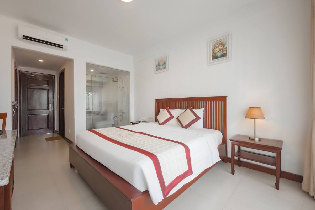 A hotel room with a large bed, a bedside table with a lamp, a shower area, and a small desk.