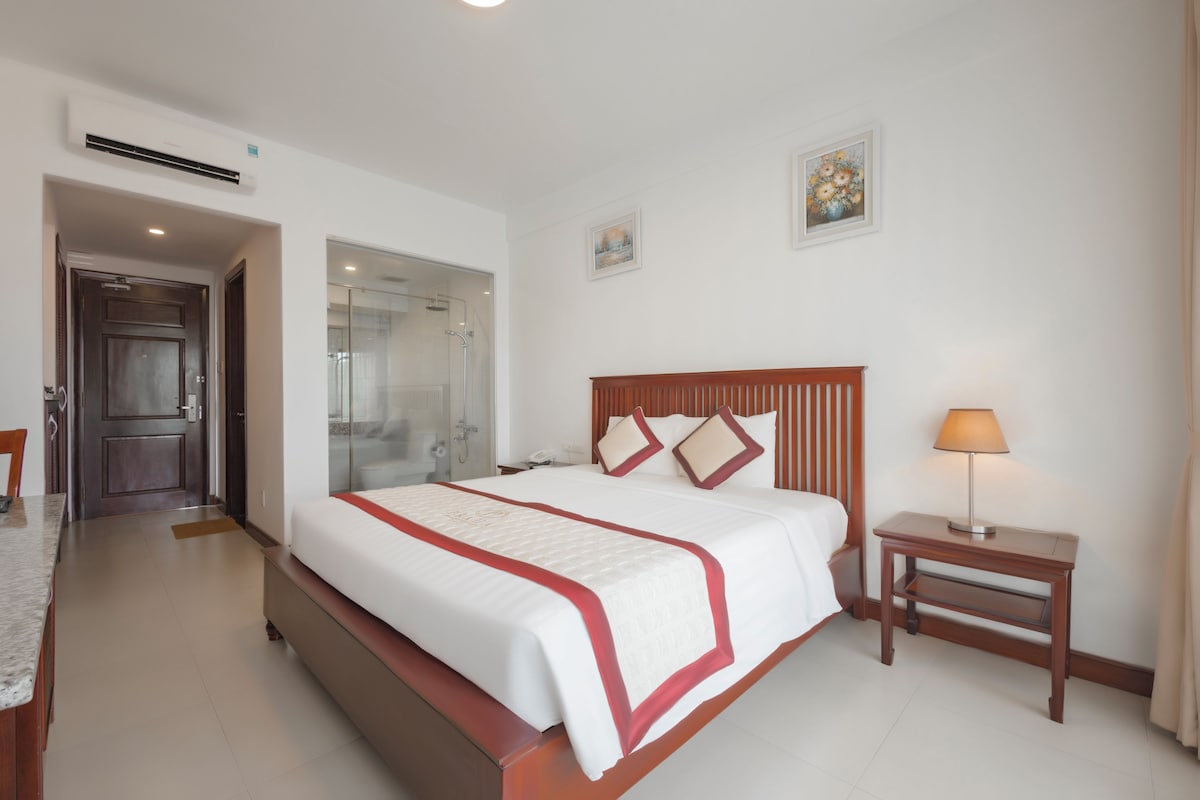 Habitación de hotel con una cama grande, una mesita de noche con lámpara, zona de ducha y un pequeño escritorio.