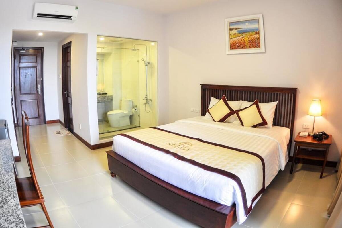 A hotel room with a large bed, a bedside table with a lamp, a bathroom with a glass shower enclosure, and a painting on the wall.