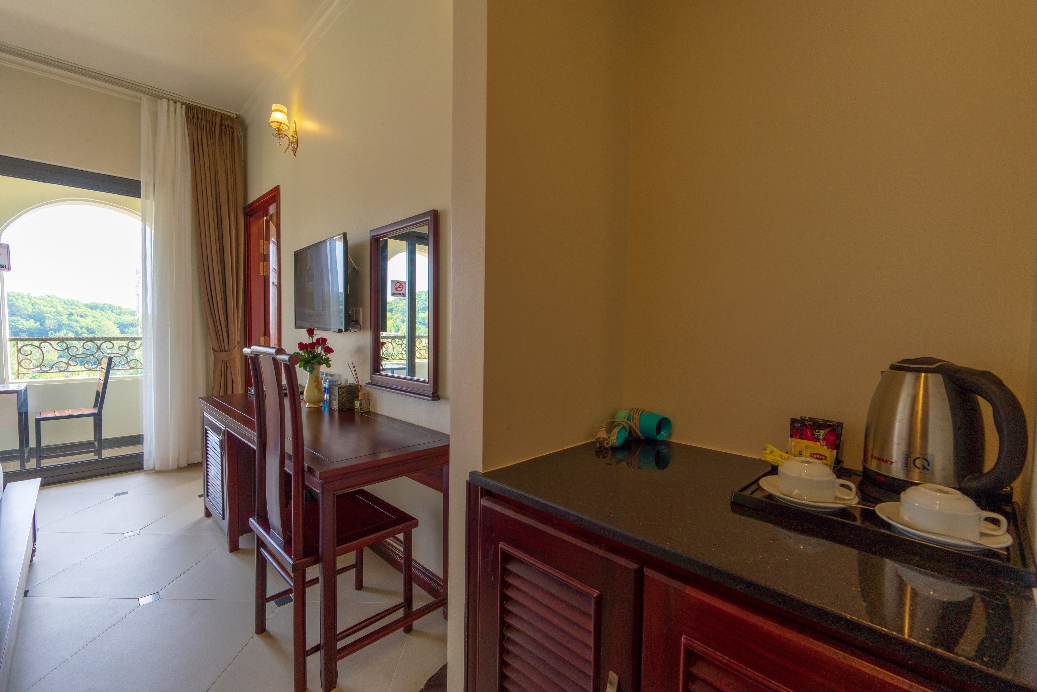 A hotel room with a dining area, a TV mounted on the wall, a window with curtains, and a kitchenette with a coffee maker and cups.