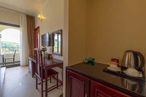 Habitación de hotel con comedor, televisión montada en la pared, ventana con cortinas y kitchenette con cafetera y tazas.