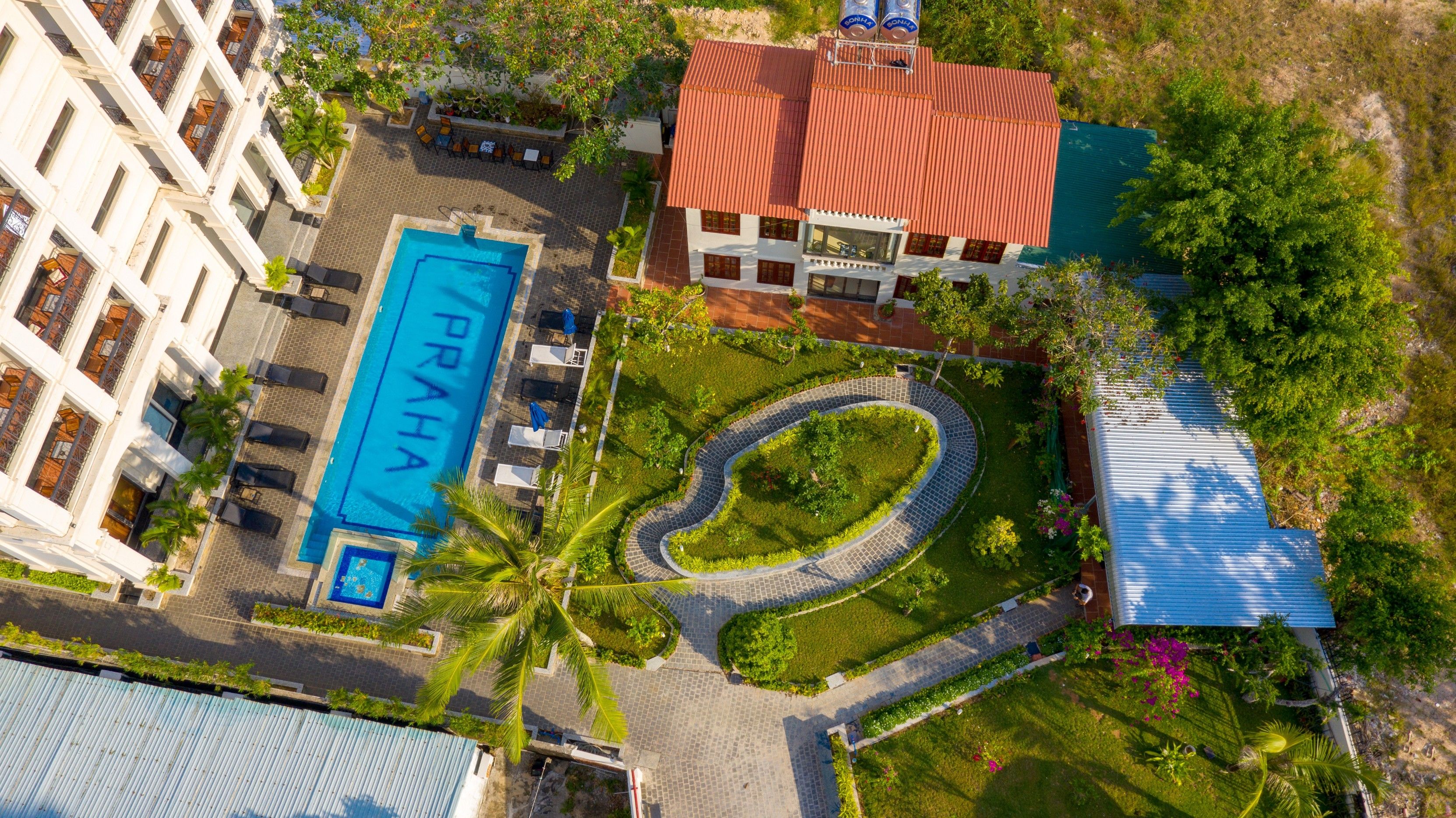 An aerial view of a hotel with a swimming pool, a landscaped garden, and a building with a red roof.