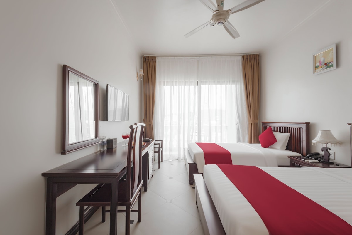 Habitación de hotel con dos camas, un escritorio, una silla, un ventilador de techo y una ventana con cortinas.