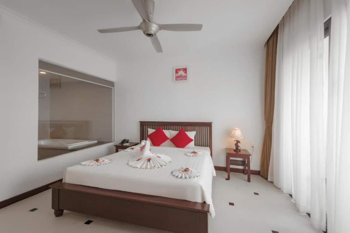 A hotel room with a bed, a ceiling fan, a bedside table with a lamp, a nightstand, and a window with curtains.