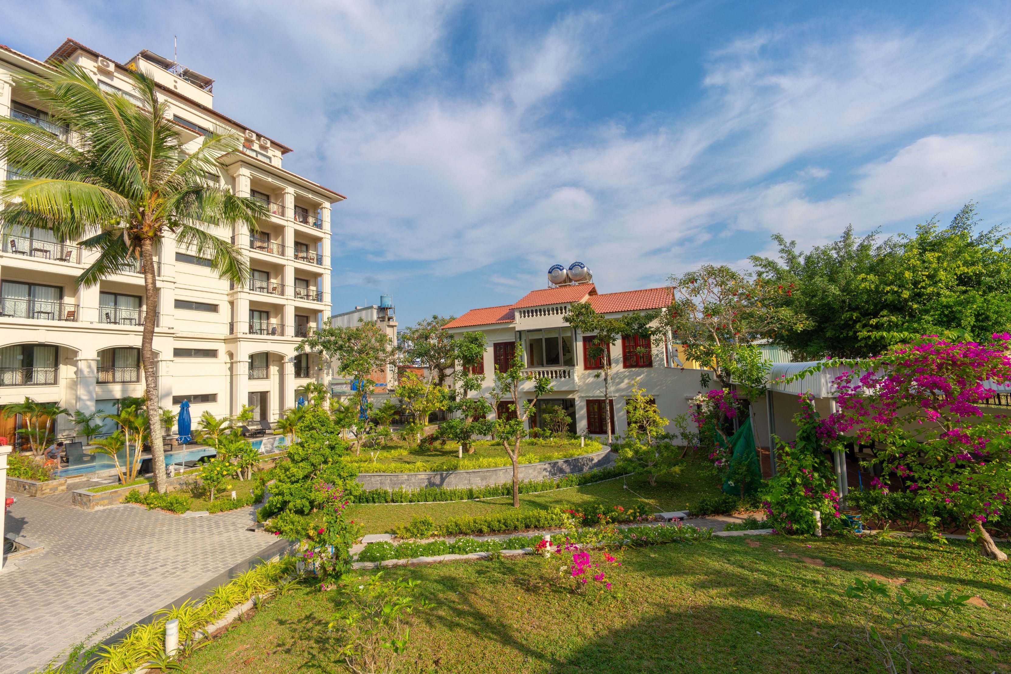 A hotel with multiple floors, outdoor swimming pool, and lush garden with various plants and flowers.