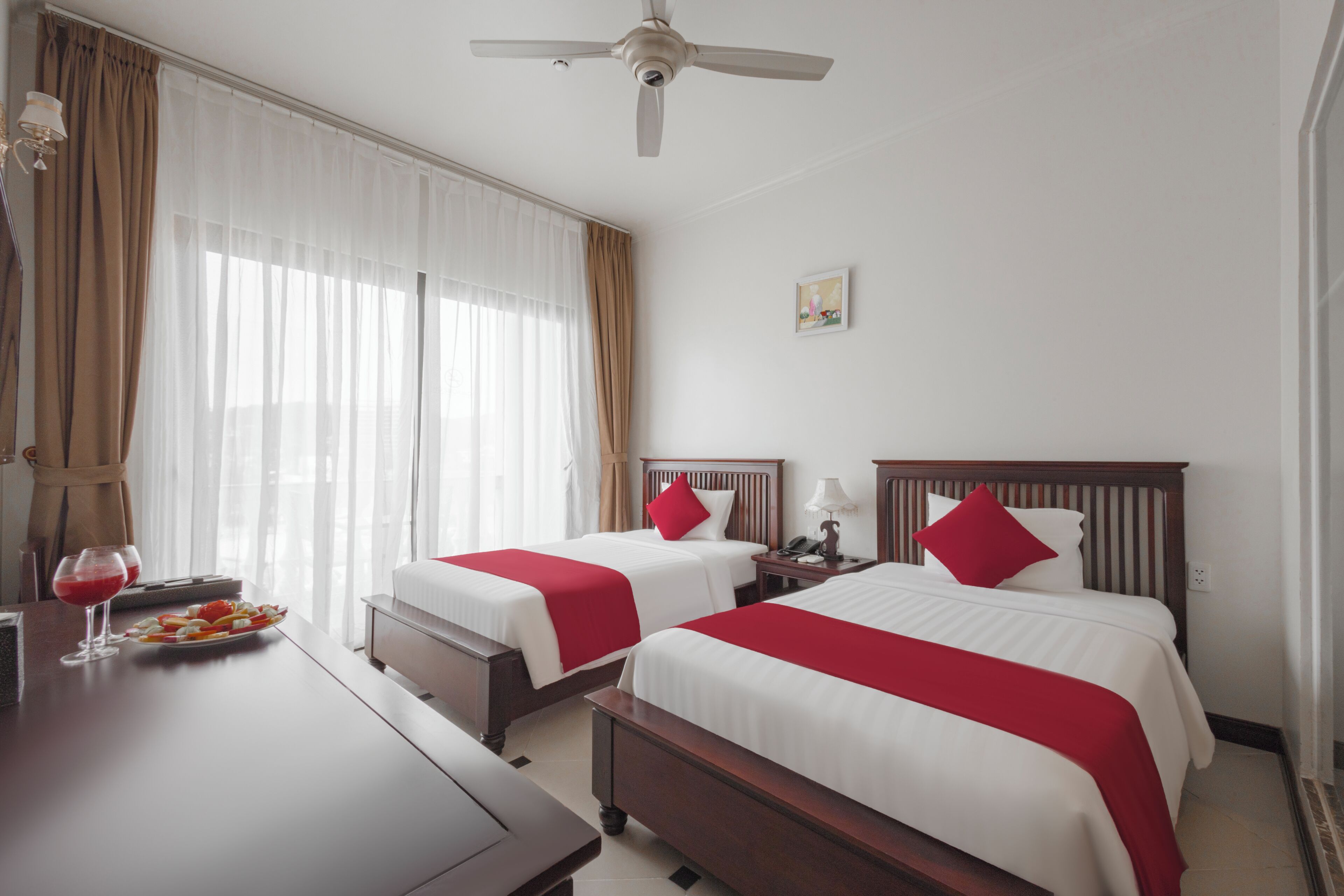 A hotel room with two beds, a ceiling fan, a bedside table with a lamp, and a window with curtains.