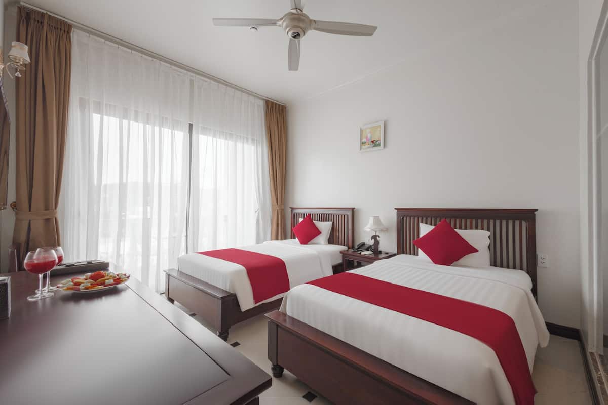A hotel room with two beds, a ceiling fan, a bedside table with a lamp, and a window with curtains.