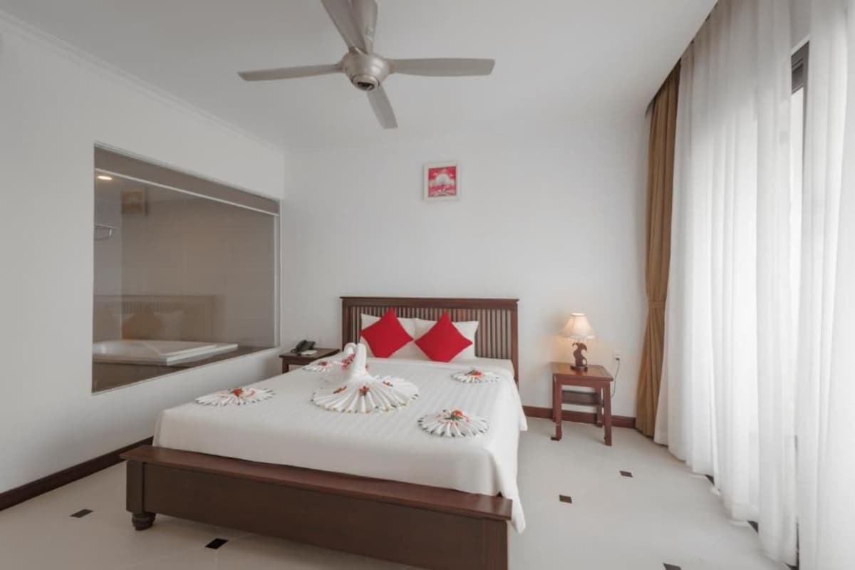 Habitación de hotel con cama, ventilador de techo, mesita de noche con lámpara, mesita auxiliar y ventana con cortinas.