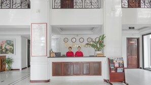 Zona de recepción de un hotel con un mostrador blanco, dos recepcionistas vestidas de rojo, un letrero de recepción, un reloj, una planta y un atril con folletos.
