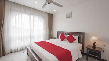 A hotel room with a bed, a bedside table with a lamp, a ceiling fan, and sheer curtains.