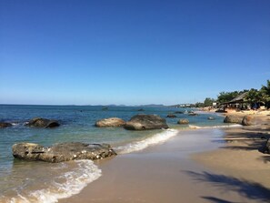 A beach with clear water, large rocks, and a distant view of a town. - Praha Hotel Phu Quoc (Phu Quoc)