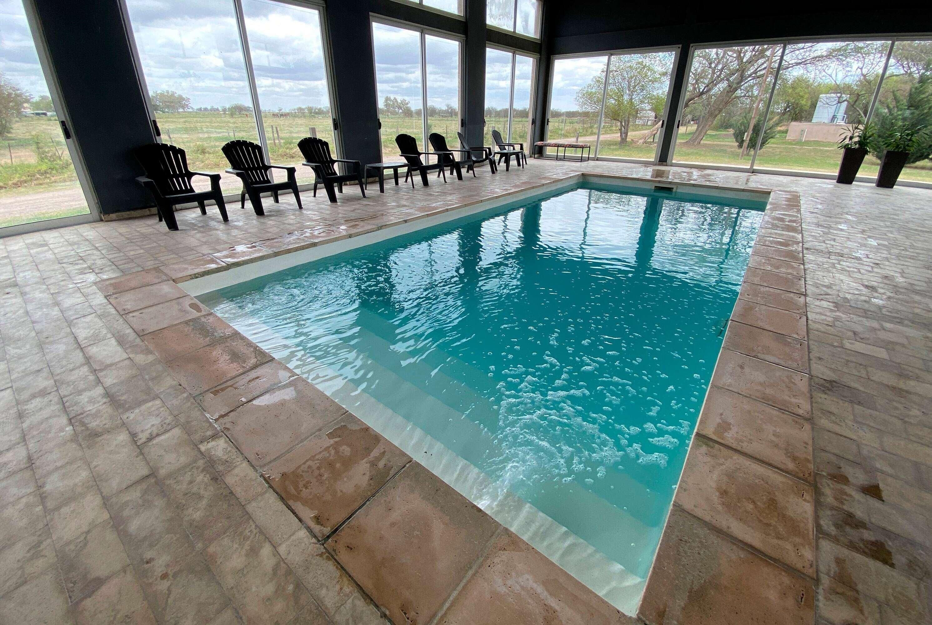 An indoor swimming pool with clear blue water, surrounded by black lounge chairs and large windows.