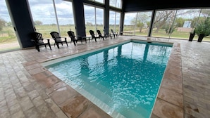 An indoor swimming pool with clear blue water, surrounded by black lounge chairs and large windows.
